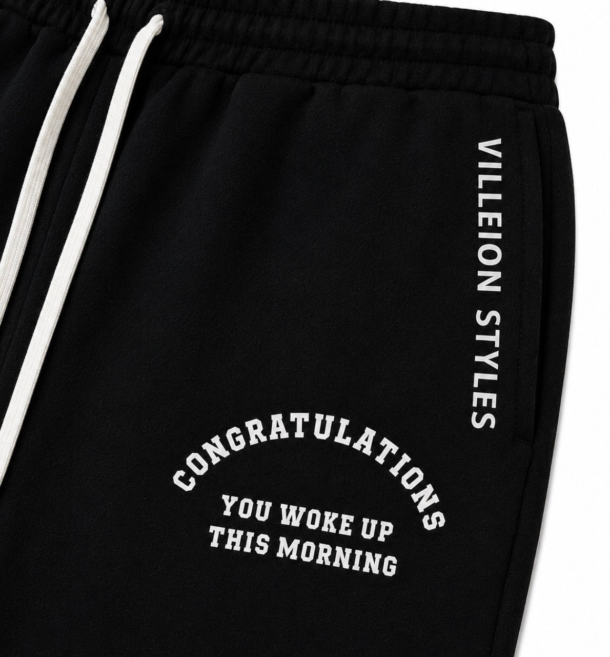 Close-up of black sweatpants with white drawstrings, white vertical text reading 'VILLEION STYLES' on the side, and white arched text reading 'CONGRATULATIONS YOU WOKE UP THIS MORNING' on the front.