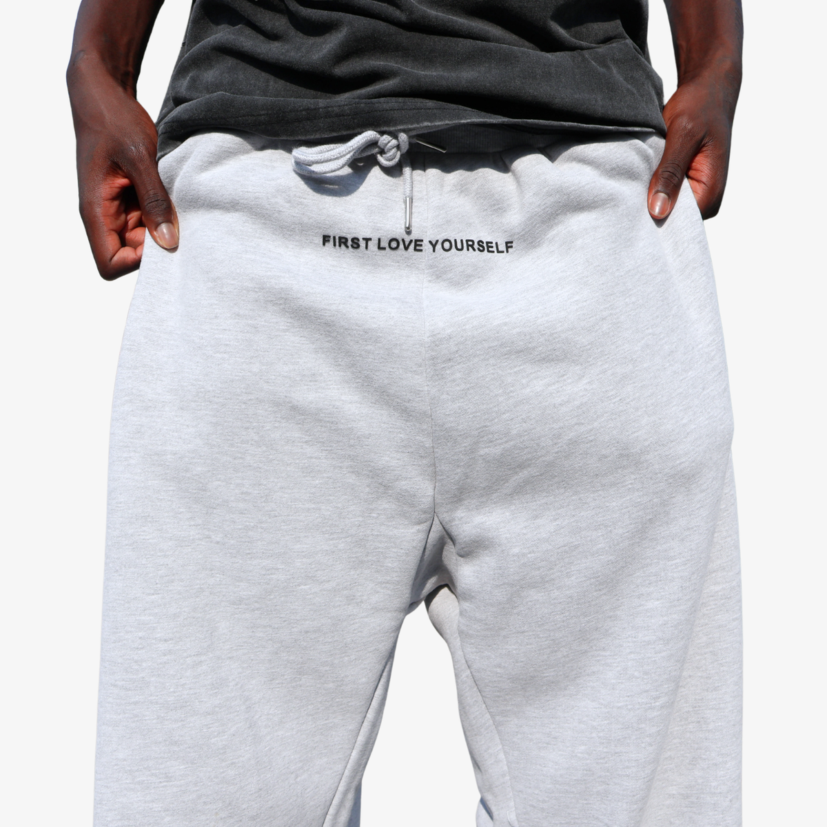 A close-up of a person wearing light gray heather sweatpants with the text 'FIRST LOVE YOURSELF' printed in black on the front waistband, paired with a dark gray t-shirt against a plain light background.